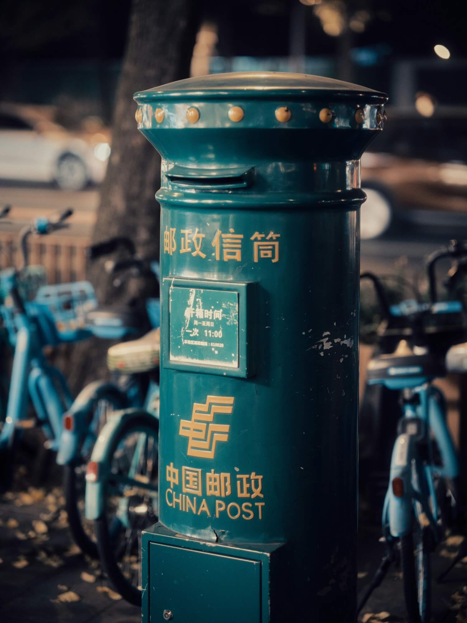 Dunkelgrüner China Post Briefkasten auf einer Straße, dahinter geparkte Fahrräder.