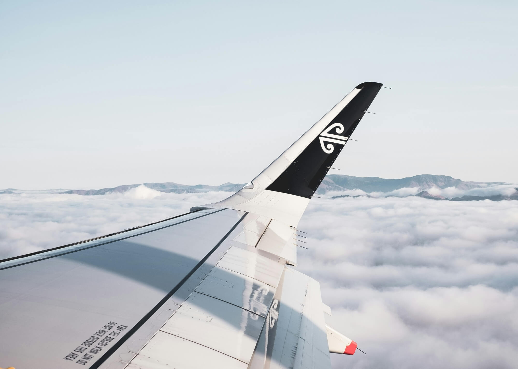 Blick aus dem Flugzeugfenster auf die Wolken und die neuseeländische Landschaft unter einem strahlend blauen Himmel.